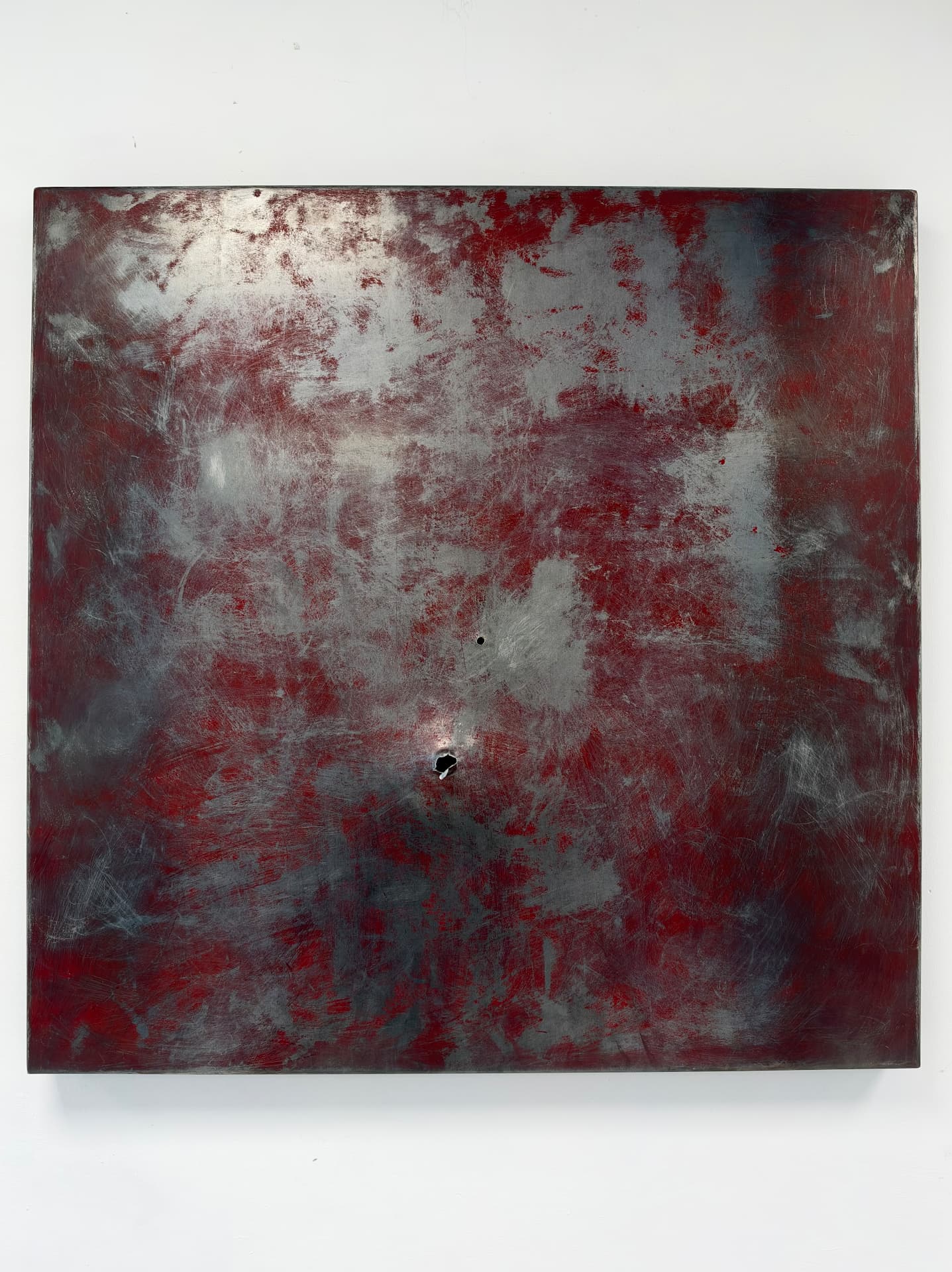 Square steel panel scuffed to bare metal across a deep red surface, a small hole and a jagged puncture below centre